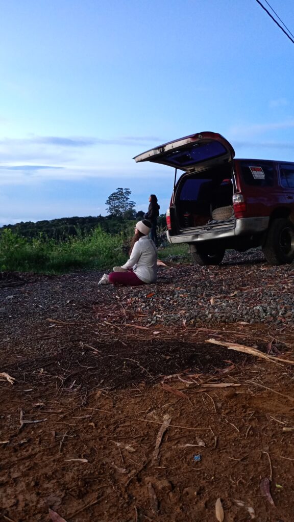 Traveler sitting on the ground beside a parked vehicle, enjoying the serene natural landscape at dusk.